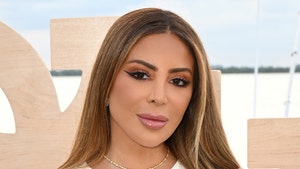 Larsa Pippen is holding part of her hair while posing for a picture in front of a wood display. She's wearing a white dress and a single necklace.