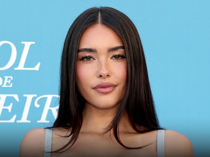 Madison Beer is seen posing for a photo in front of a blue wall. She's wearing a dress with light blue straps.