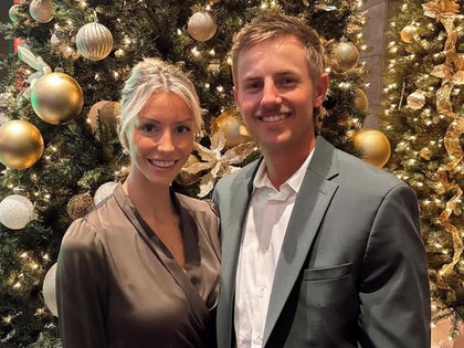 Makena White and Jake Knapp are seen at what appears to be a holiday party. She's wearing a brown dress, while he's wearing a gray coat and a white shirt.