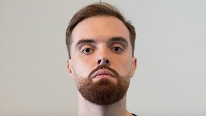 A man wearing a black t-shirt and standing in front of a white wall is staring directly at the camera.