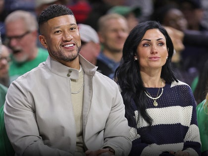 Marcus and Joanna Freeman are sitting next to each other at what appears to be a sporting event. He's wearing a beige jacket and an off-white sweater, while she's wearing a blue-and-white striped pullover.
