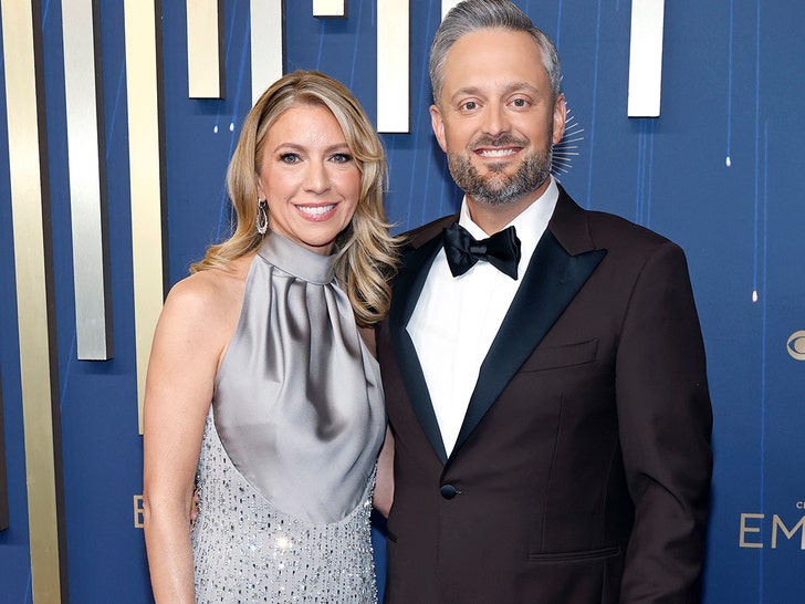 Laura Bargatze is seen while posing next to her husband Nate for a photo. She's wearing a sparkling dress with a light gray upper portion. He's wearing a brown jacket, white shirt, and black bowtie.
