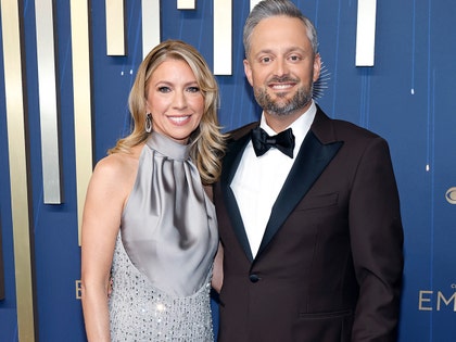 Laura Bargatze is seen while posing next to her husband Nate for a photo. She's wearing a sparkling dress with a light gray upper portion. He's wearing a brown jacket, white shirt, and black bowtie.