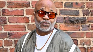 Damon Dash is standing in front of a brick wall. He's wearing a white t-shirt, gray puffer jacket, and orange-tinted sunglasses.