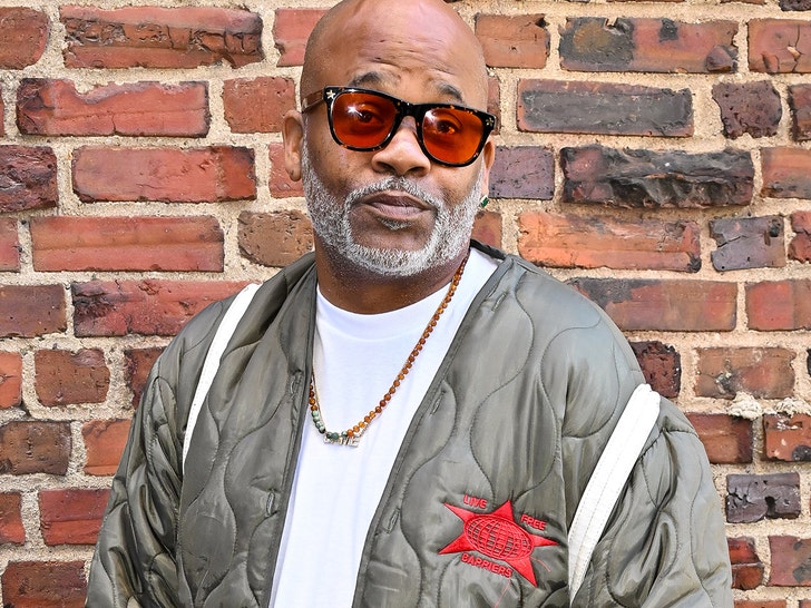 Damon Dash is standing in front of a brick wall. He's wearing a white t-shirt, gray puffer jacket, and orange-tinted sunglasses.