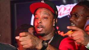 21 Savage is seen pointing at an offscreen object. He's wearing a black shirt with red sleeves, as well as a tri-colored hat.