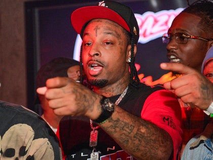 21 Savage is seen pointing at an offscreen object. He's wearing a black shirt with red sleeves, as well as a tri-colored hat.