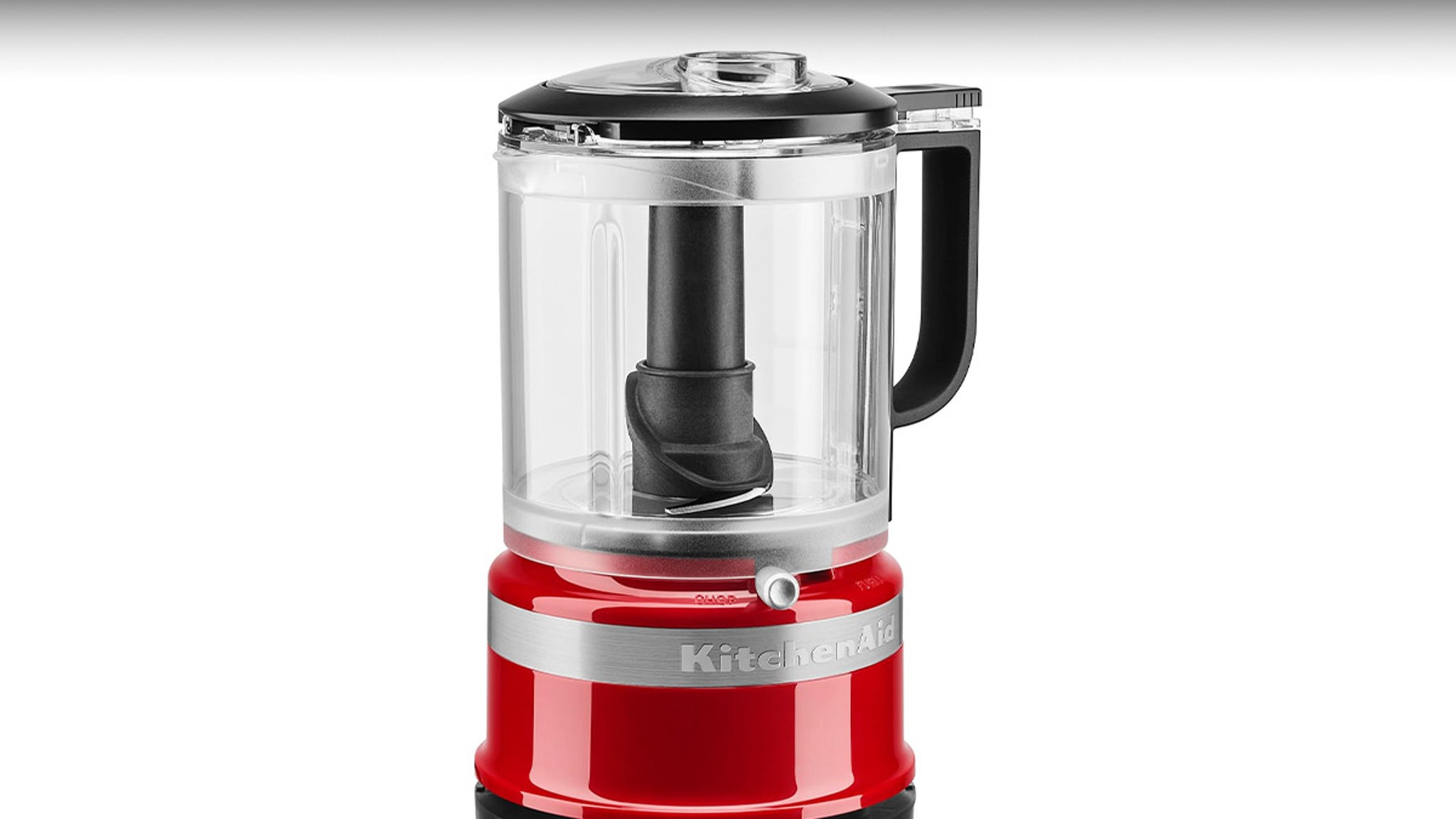 The image shows a KitchenAid food processor with a glossy red base and a clear plastic container on top. The container has a black handle and a black central blade assembly inside. A silver band around the base displays the KitchenAid brand name, and the lid is black with a small transparent section.