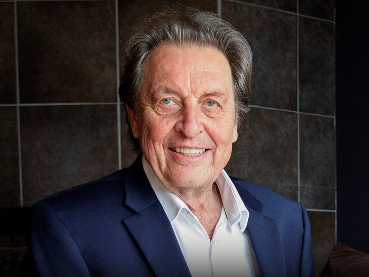 Errol Musk is smiling while standing in front of a tile wall. He's wearing a white shirt and a navy blue jacket.