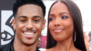 Keith Powers and Ryan Destiny are seen smiling next to each other. He's wearing a black shirt and a silver necklace, while she's wearing a white dress.