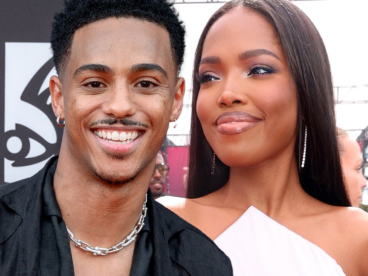 Keith Powers and Ryan Destiny are seen smiling next to each other. He's wearing a black shirt and a silver necklace, while she's wearing a white dress.