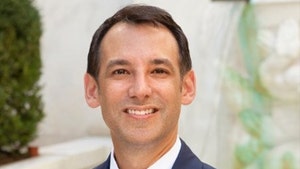 A man wearing a navy jacket, white shirt and light purple tie is smiling while standing in a courtyard.