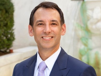 A man wearing a navy jacket, white shirt and light purple tie is smiling while standing in a courtyard.
