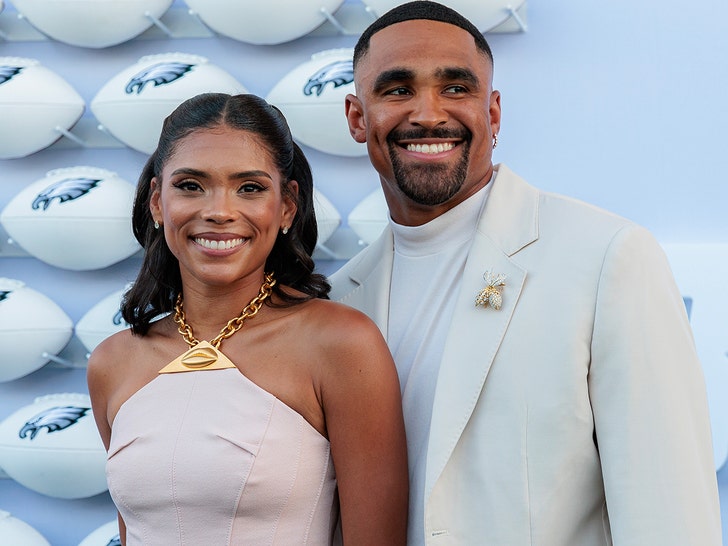 Bry Burrows is pictured standing next to her husband Jalen Hurts. She's wearing a beige dress with a gold necklace, and he's wearing an off-white jacket and a cream-colored turtleneck sweater.