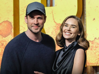 Liam Hemsworth and Gabriella Brooks are seen at a movie premiere. He's wearing a dark blue crewneck sweater and she's wearing a black dress.