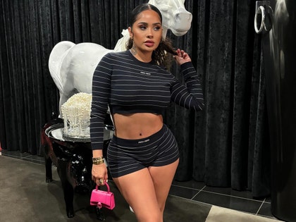 Aileen Lopera is seen standing in front of a white plaster horse, black curtains, and a wood table. She's wearing a striped black long-sleeve top that exposes her midsection, as well as matching shorts. She's accessorized with various pieces of jewelry and a small hot pink purse.