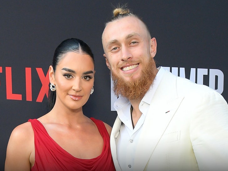 George Kittle is smiling while standing next to his wife, Claire, at an even for Netflix. He's wearing a cream-colored jacket and a white shirt, while she's wearing a red dress.