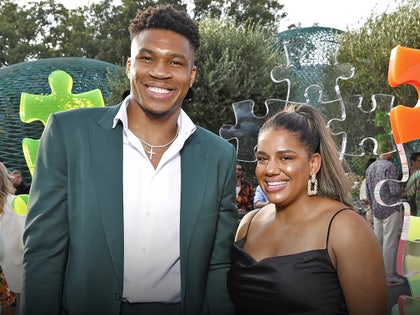 Giannis Antetokounmpo and his Mariah Riddlesprigger are seen posing in front of several oversized puzzle pieces. He's wearing a dark green jacket and a white shirt, while she's wearing a black dress.