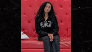 Celeste Rivas is seen sitting on a large red couch. She's wearing a Gap sweater, sweat pants, and black-and-white Vans.