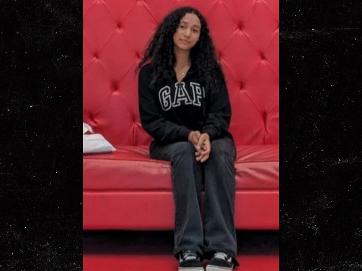 Celeste Rivas is seen sitting on a large red couch. She's wearing a Gap sweater, sweat pants, and black-and-white Vans.