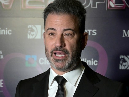 Jimmy Kimmel is seen at a red carpet event. He's wearing a black jacket, a matching tie, and a white button-up shirt.