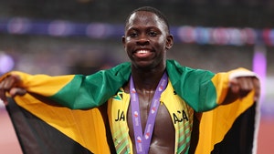 Oblique Seville is holding his arms out while wearing a gold medal on top of an unzipped running outfit. He's got the Jamaican flag draped over his shoulders.