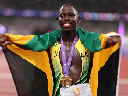 Oblique Seville is holding his arms out while wearing a gold medal on top of an unzipped running outfit. He's got the Jamaican flag draped over his shoulders.