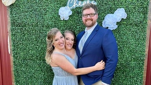 Abby and Brittany Hensel are hugging Abby's husband Josh Bowling. She's wearing a blue dress, and he's wearing a blue jacket and khakis. They're standing in front of a grass wall panel with fake flowers and a neon sign reading 