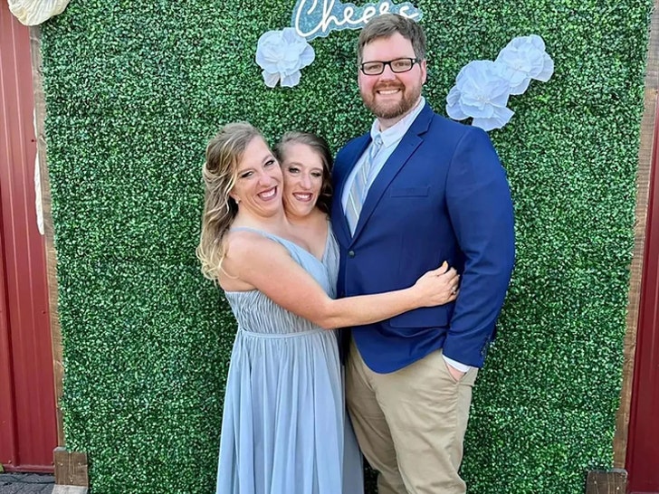 Abby and Brittany Hensel are hugging Abby's husband Josh Bowling. She's wearing a blue dress, and he's wearing a blue jacket and khakis. They're standing in front of a grass wall panel with fake flowers and a neon sign reading 