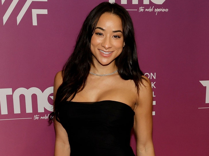 A woman wearing a black dress and silver necklace is standing in front of a burgundy wall featuring several logos.