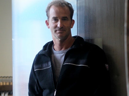 Andrew Cabot is seen while posing in front of a wood door. He's wearing a gray t-shirt underneath a black hoodie.
