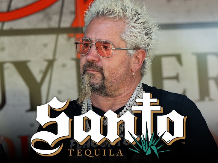 Guy Fieri's looking concerned behind the logo for Santo Tequila. He's wearing a black t-shirt, several chains, and orange-tinted aviator-style sunglasses.