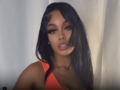 Tomeka Thiam is seen posing in front of what appears to be white drapes. She's wearing a red-orange dress.