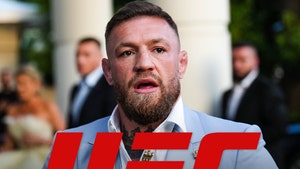 Conor McGregor is looking concerned behind the logo of the UFC. He's wearing a light blue jacket and a white button-up shirt.