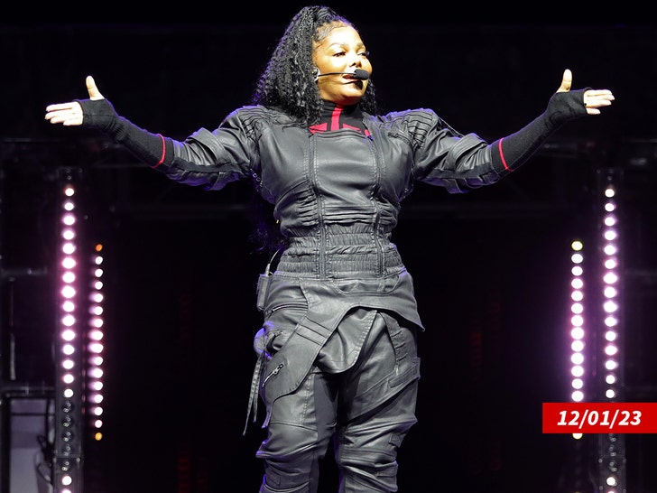 Janet Jackson wearing a one-piece black outfit with leather-like material and a microphone in front of her mouth.