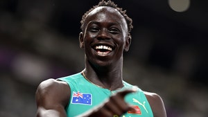 Gout Gout is seen smiling and pointing at a sports event. He's wearing a turquoise running top featuring the Australian flag and the logo for Puma.