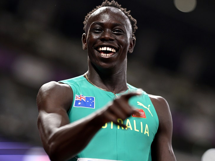 Gout Gout is seen smiling and pointing at a sports event. He's wearing a turquoise running top featuring the Australian flag and the logo for Puma.