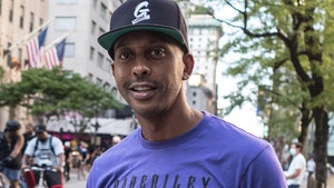 Gillie Da Kid's seen while standing on a busy street corner. He's wearing a purple t-shirt and a black baseball hat.