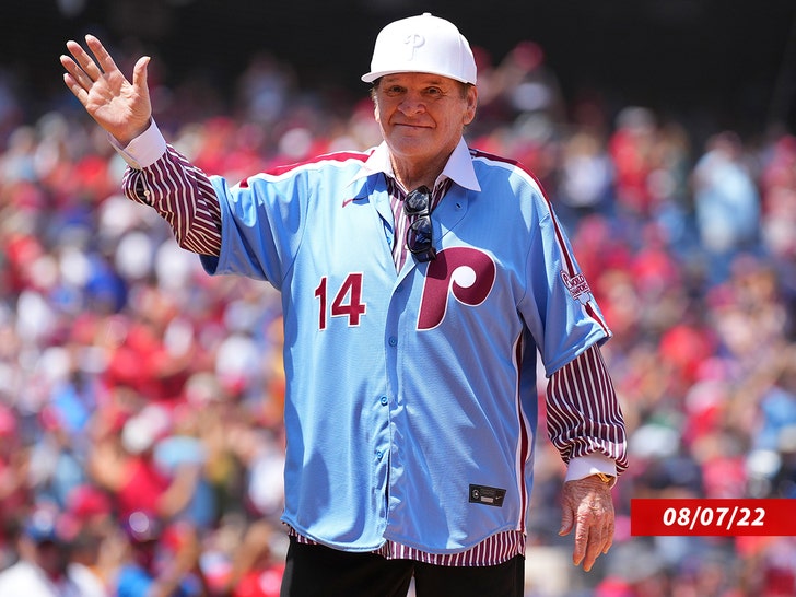 Pete Rose acenando enquanto usava uma camisa listrada sob uma camisa azul do Philadelphia Phillies e um boné branco dos Phillies.