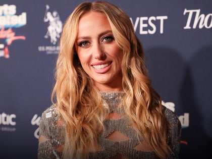 A woman wearing a sparkling silver dress is smiling while posing in front of a navy blue wall with several logos running across its surface.