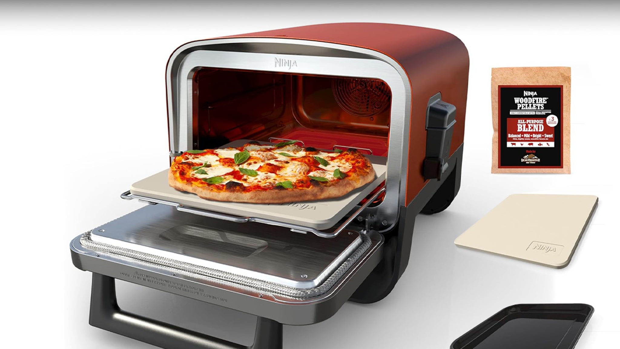 The image shows a compact countertop pizza oven with a pizza stone slide-out tray, on which a freshly baked pizza with melted cheese, tomato sauce, and fresh basil leaves rests. The oven has a rounded rectangular shape with a reddish-brown exterior and a metallic silver front frame branded 