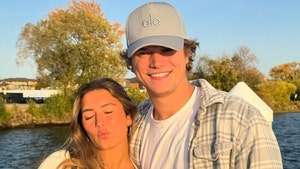 J.J. McCarthy and Katya Kuropas are standing in front o fa lake. She's wearing a white dress and an off-white sweater. He's dressed in a white shirt, gray flannel and an Alo Yoga cap.
