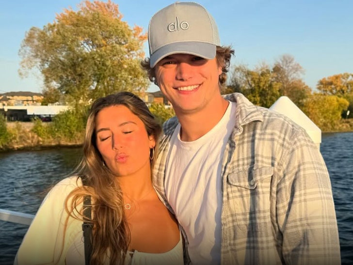 J.J. McCarthy and Katya Kuropas are standing in front o fa lake. She's wearing a white dress and an off-white sweater. He's dressed in a white shirt, gray flannel and an Alo Yoga cap.