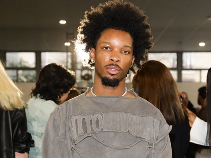 D4vd is seen posing for a photo at an event. He's wearing a distressed gray shirt and a necklace.