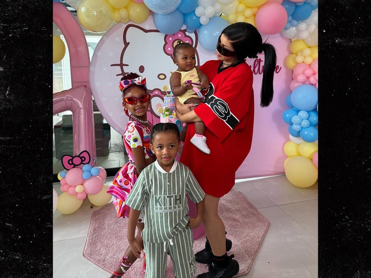 Cardi B is seen while posing with her kids Kulture, Wave, and Blossom, in front of a Hello Kitty-themed balloon display. She's wearing an oversized red shirt and what appears to be a pair of wraparound sunglasses.