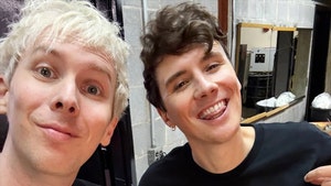 YouTubers Dan and Phil are seen posing for a selfie. They're both wearing black shirts, and Dan's shirt features text reading 
