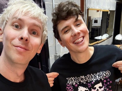 YouTubers Dan and Phil are seen posing for a selfie. They're both wearing black shirts, and Dan's shirt features text reading 