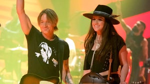 Keith Urban and Maggie Baugh are seen at a concert. He's wearing a black shirt and matching pants, and he has a Telecaster-style guitar slung over his left shoulder. She's wearing a black shirt with a matching vest and pants, as well as a wide-brimmed hat. She also has a mandolin slung over her left shoulder.