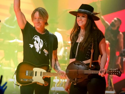 Keith Urban and Maggie Baugh are seen at a concert. He's wearing a black shirt and matching pants, and he has a Telecaster-style guitar slung over his left shoulder. She's wearing a black shirt with a matching vest and pants, as well as a wide-brimmed hat. She also has a mandolin slung over her left shoulder.