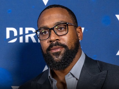 Marcus Jordan is pictured in front of a graphic for DirecTV. He's wearing a dark blue jacket on top of a light gray shirt.